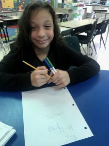 Student sits a at blue desk smiling, while writing a piece of lined notebook paper.