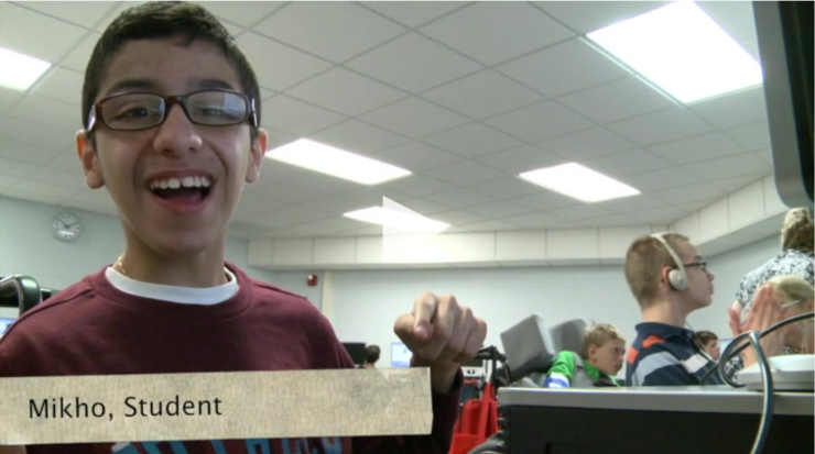 A student named Mikho smiles at the camera in a classroom with other students working in the background.