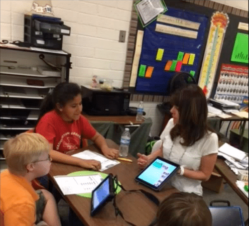 Teacher engages in a word study with three students at a desk, in a classroom.