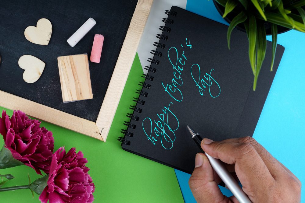 Depositphotos292932604s2019 Building Wings Building Wings A hands writes Happy Teachers Day in turquoise ink on a black note pad on a blue and green surface