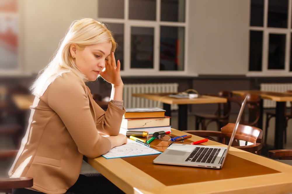 Depositphotos322248566s2019 Building Wings Building Wings An adult sitting at the desk in the front of an empty classroom looks thoughtfully at her computer