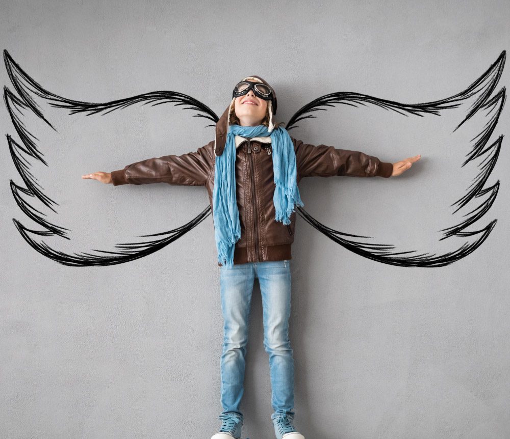 A child in a pilot outfit smiles with outstretched arms in front of large wings drawn on a grey wall