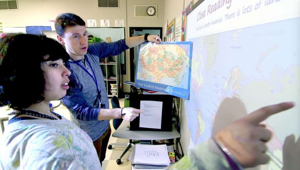 ErlindInfotextmap Building Wings Building Wings Erlind Lacy holds up a physical map of the US while a student points to a digital map of the world on a screen in a classroom