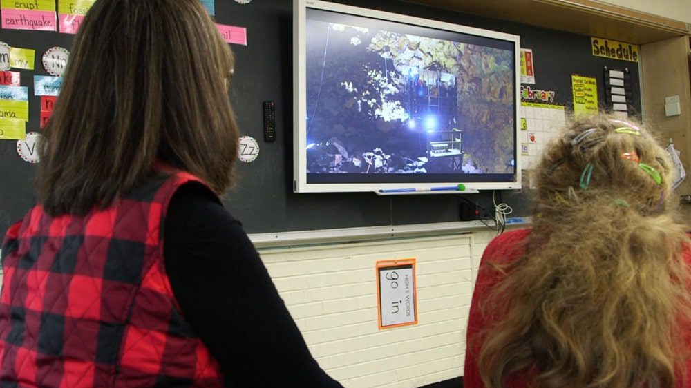 An educator wearing a red and black checkered vest sits next to a student wearing a red shirt, as they watch a geography lesson showing a map, on a smart board in a classroom.