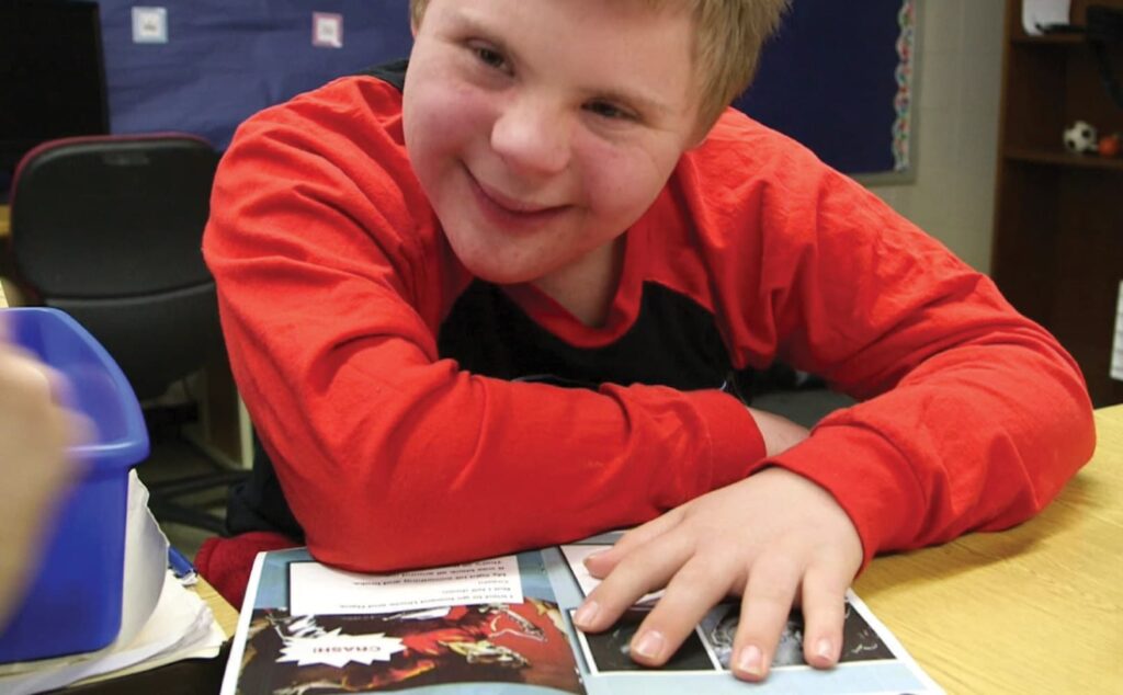 A student wearing a red shirt, smiles while reading a Readtopia graphic novel, at a desk in a classroom.