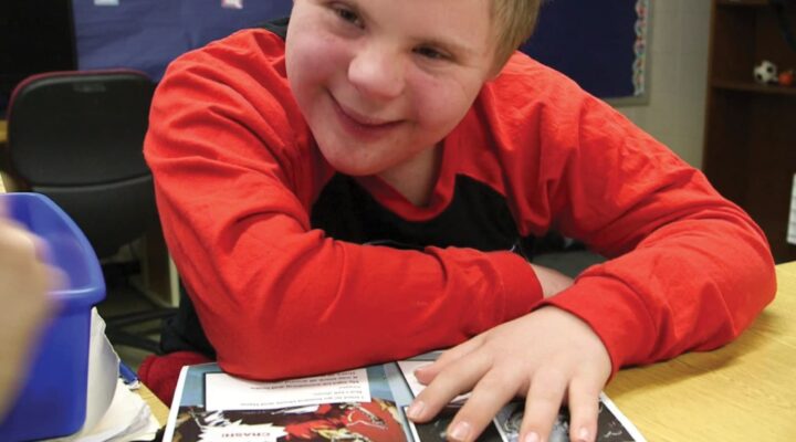 A student wearing a red shirt, smiles while reading a Readtopia graphic novel, at a desk in a classroom.