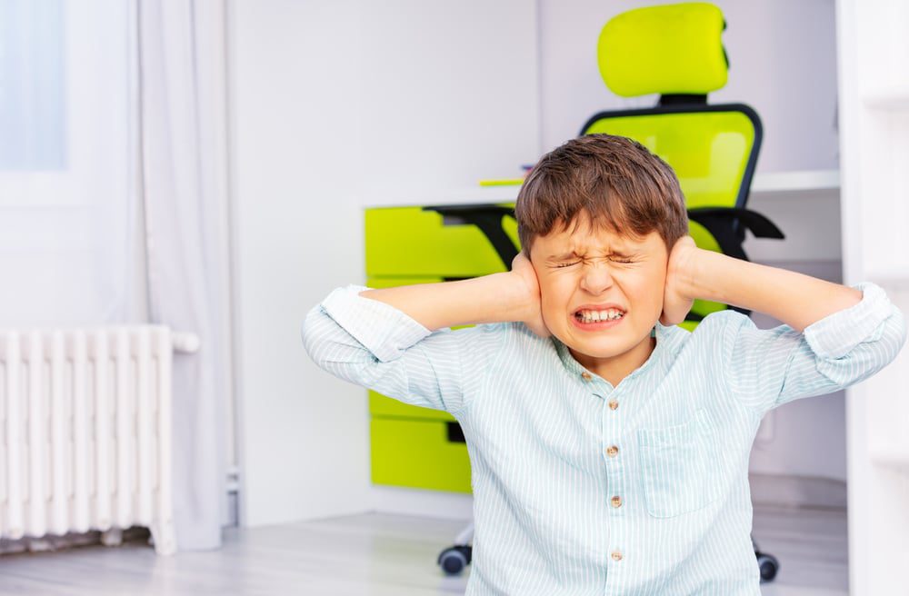 Depositphotos_410470288_S A young child holds his hands to his ears with an uncomfortable look on his face and his eyes closed.