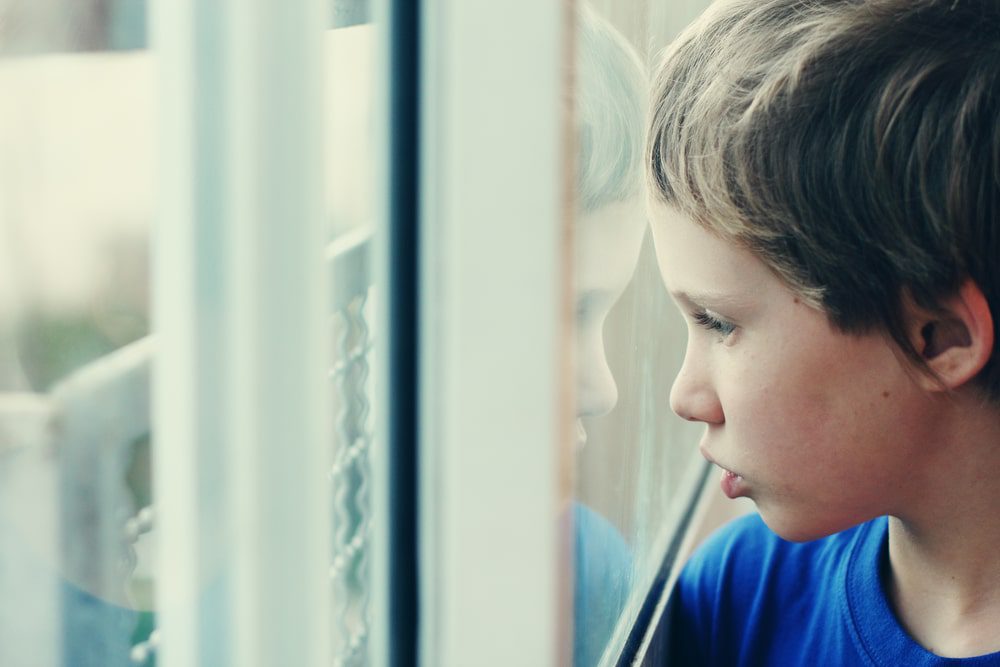 A young child in a blue shirt stares longingly out a window.