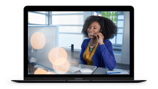 A laptop screen displays a technical support worker with a headset on smiling while working at a desk