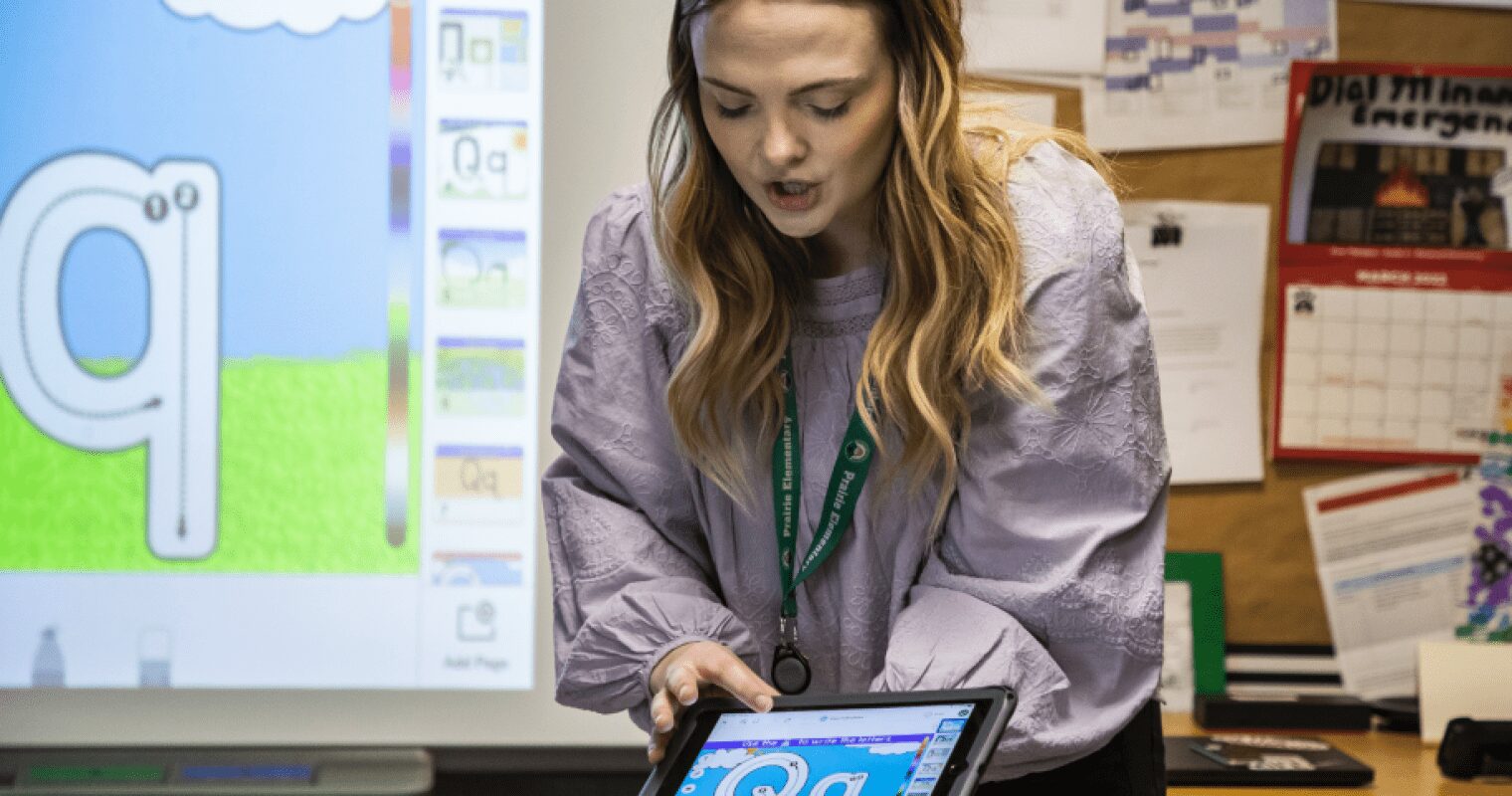 An educator points to the letter Q on an AAC device, while the same screen is presented on a smart board behind her, in a classroom.
