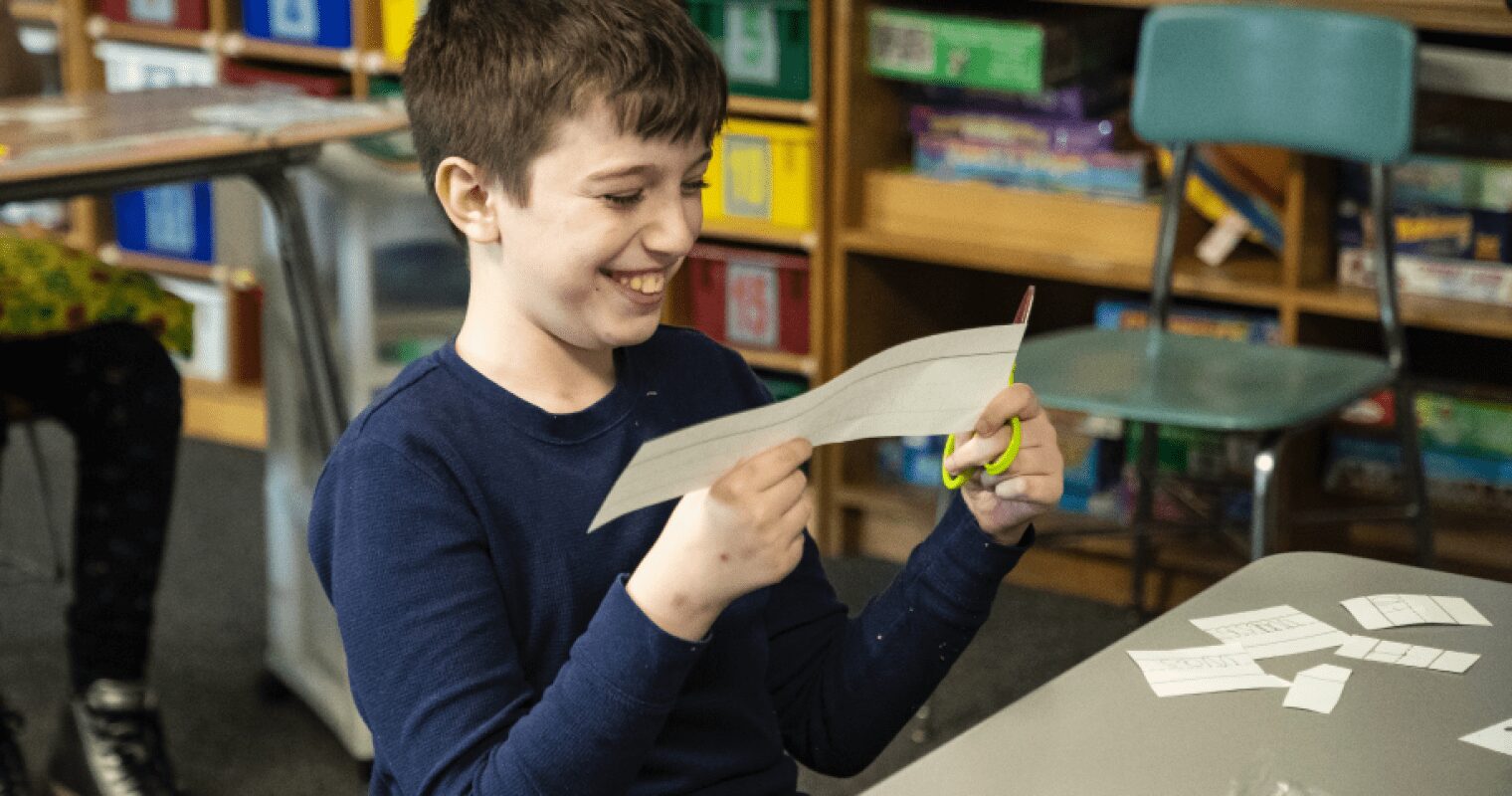 A student smiles while making cut outs of words on paper, at a desk in a classroom.