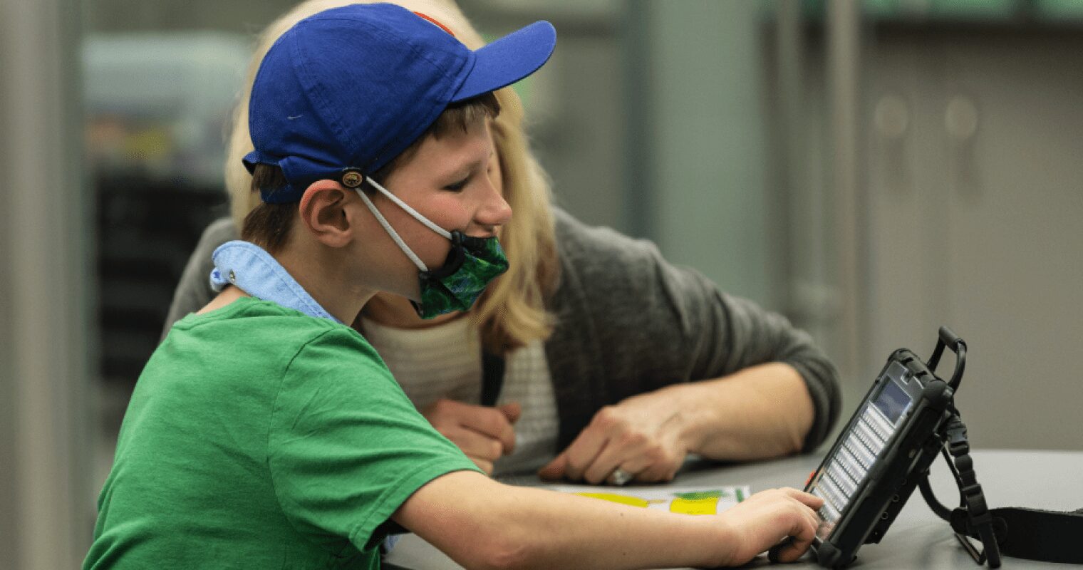 A student wearing a green shirt, blue hat, and green mask, types on an AAC device at a desk with a teacher watching next to him.