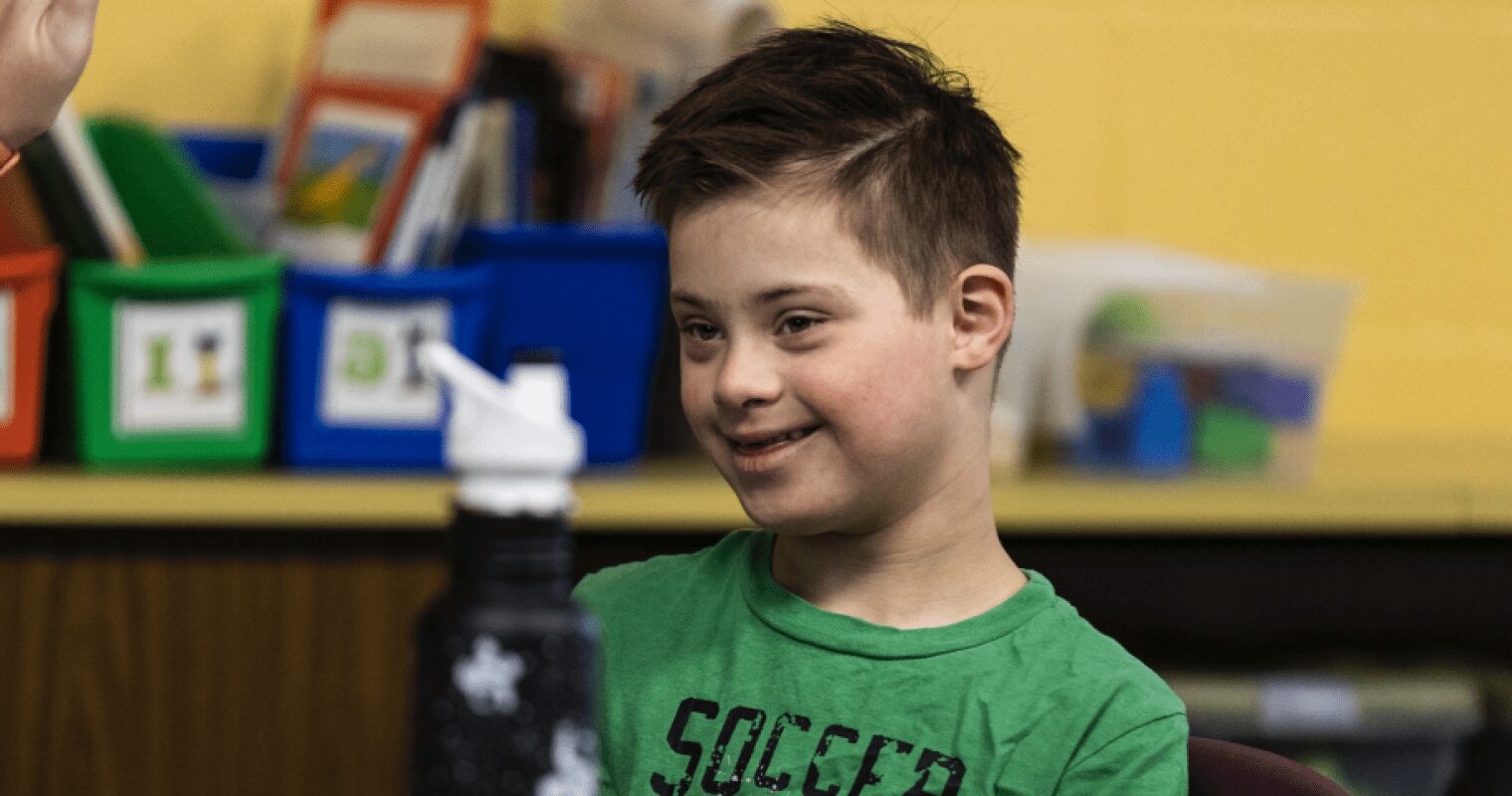 A young student in a green shirt smiles at something off camera in a classroom.