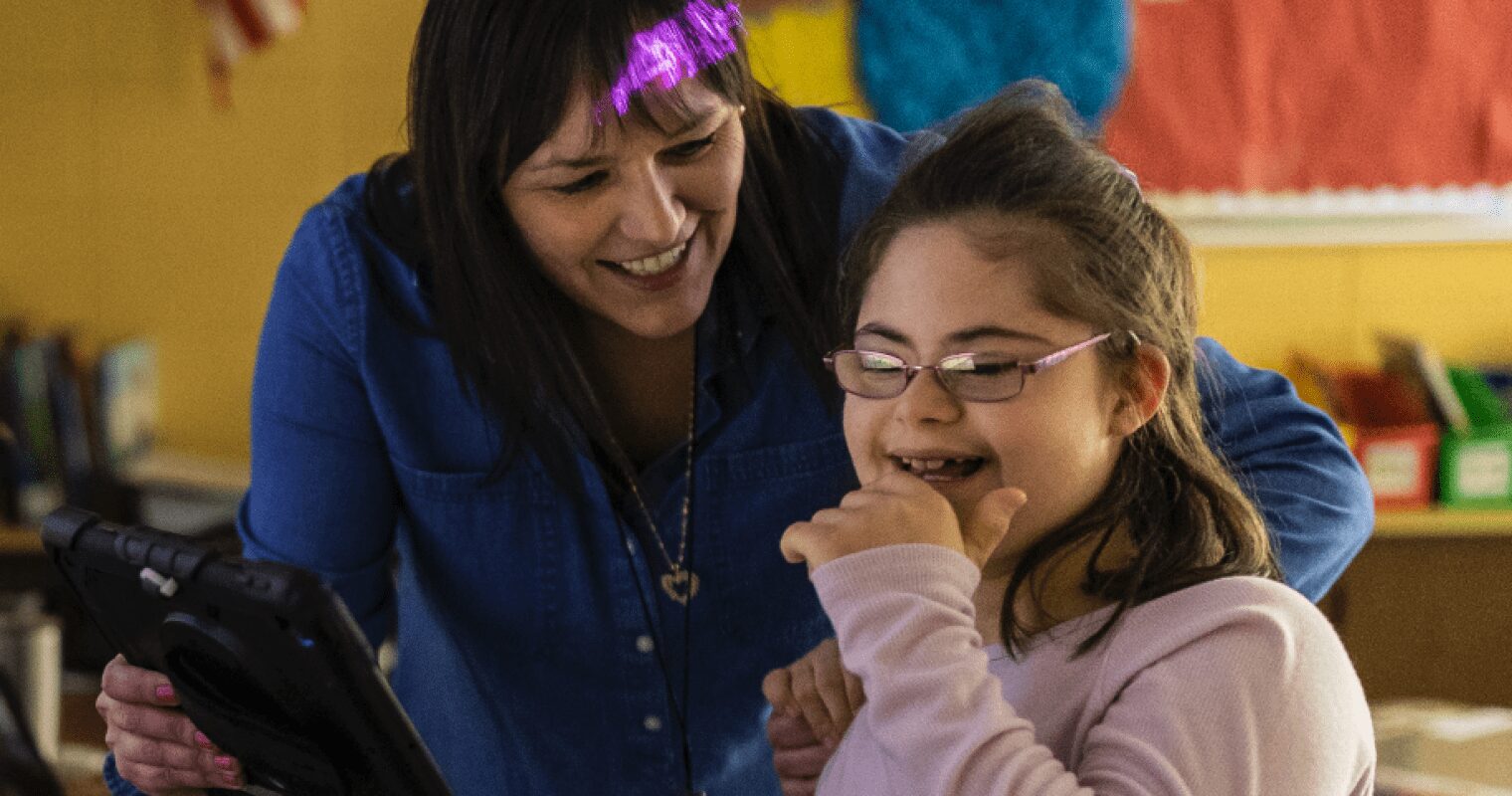 An educator and a student laughs together in a classroom, as the teacher holds up an AAC device.