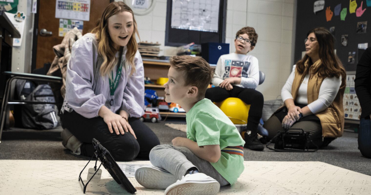 An educator plays with a student using an AAC device in a classroom and watching something on a screen at the front of the room. Another teacher and student, sitting on a ball chair, watch in the background.