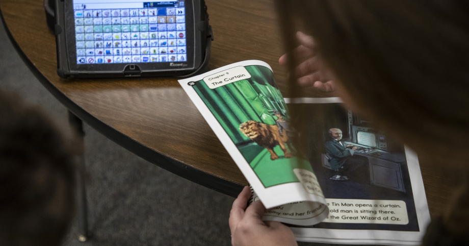 A teacher and student flip through Readtopia's "The Wizard of Oz" graphic novel, at a desk with an AAC device on in the background.