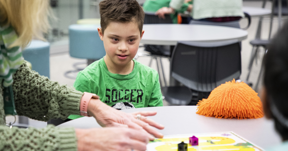 A young student, wearing a green shirt, watches a teacher work on an educational activity at a desk, in a classroom. An orange squish toy sits on the desk next to the student.