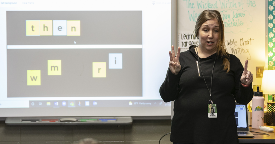 An educator in a classroom, wearing a black shirt, holds up three fingers on one hand and one finger on the other, while presenting a word lesson from a smart board.