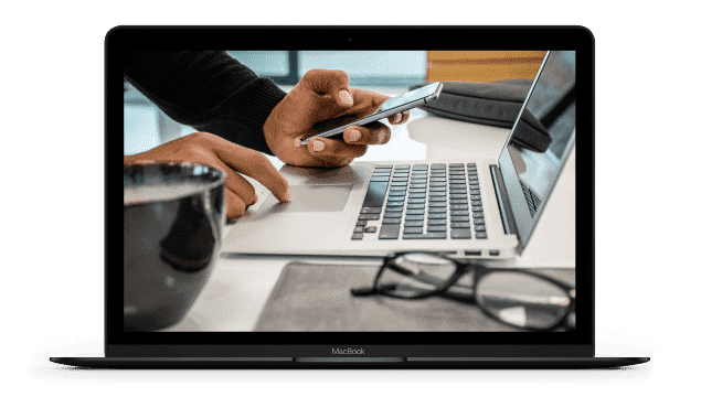 A blogger uses their smart phone and laptop while sitting at a desk with a cup of coffee On the desk there is a pair of reading glasses and a laptop case