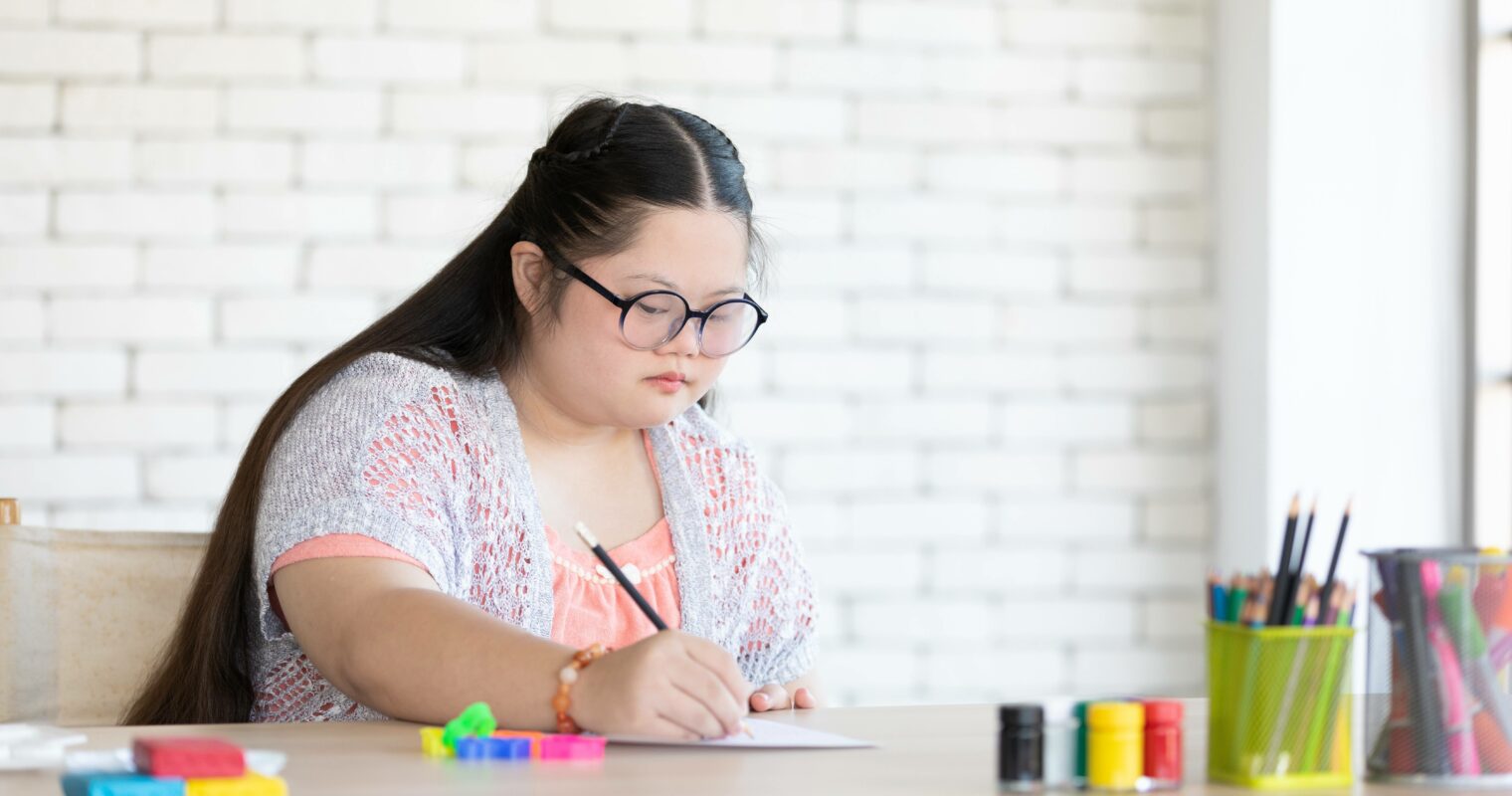 A student in a coral shirt, writes on a piece of paper at a desk, with a bright white brick background