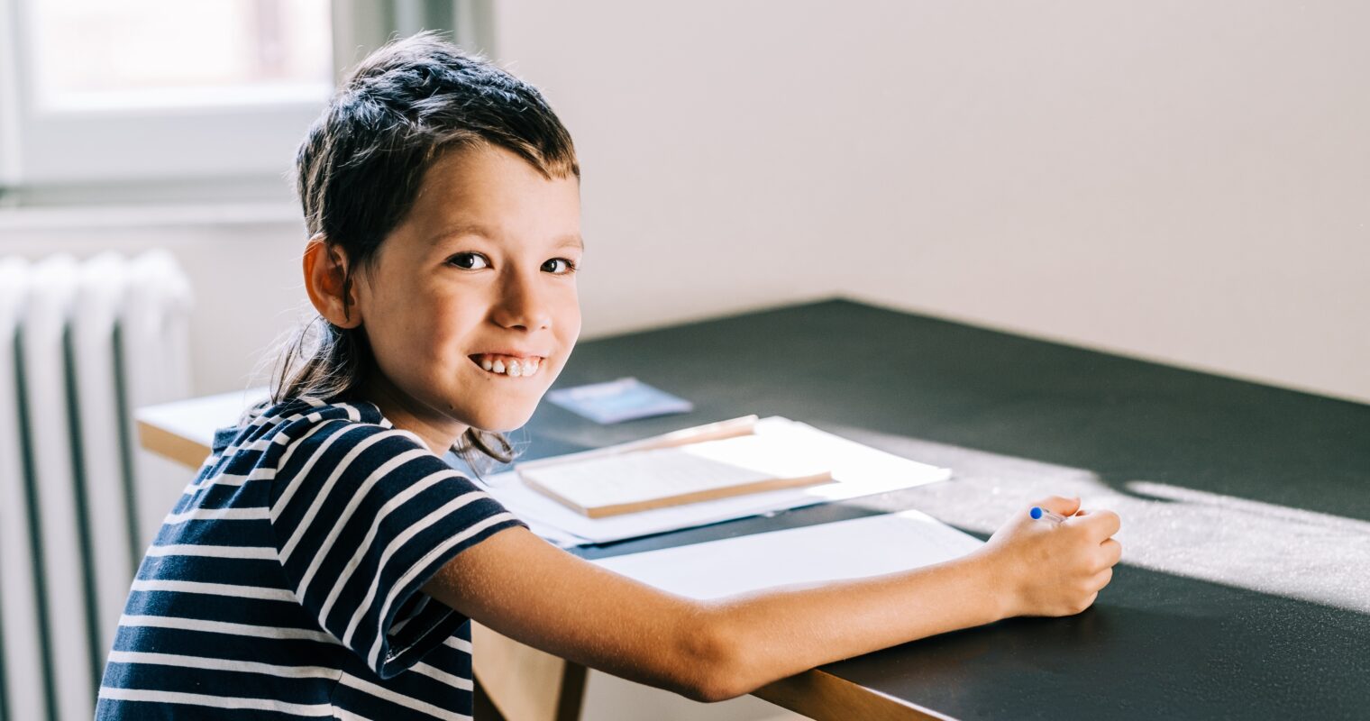A student working at a desk, wearing a black and white striped shirt, smiles at the camera with a pencil in hand.