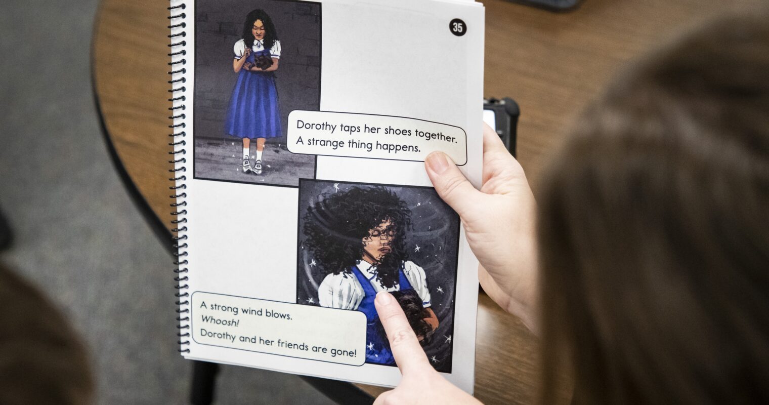 A person pointing to the page of a book in a classroom, with the book showing 2 pictures of Dorothy from The Wizard of Oz and text overlay that reads, "Dorothy taps her shoes together. A strange thing happens. A strong wind blows. Whoosh! Dorothy and her friends are gone!"