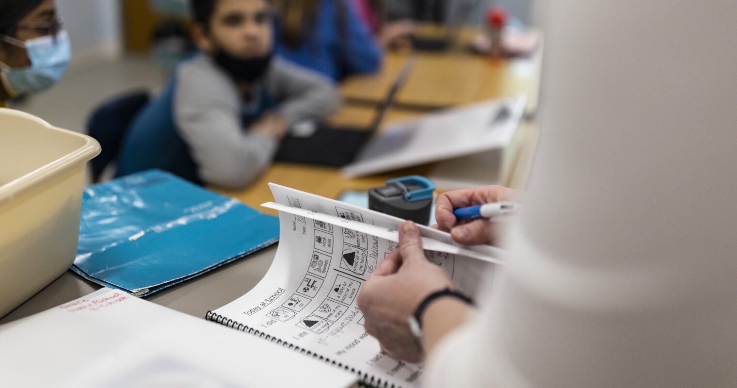 A teachers hands open a book with a student sitting at a desk blurred in the background