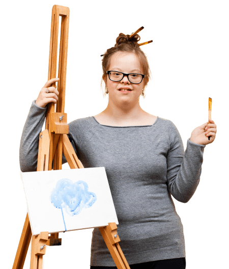 Our CommunityBanner Building Wings Building Wings Student in a grey shirt smiles by an easel with artwork on it