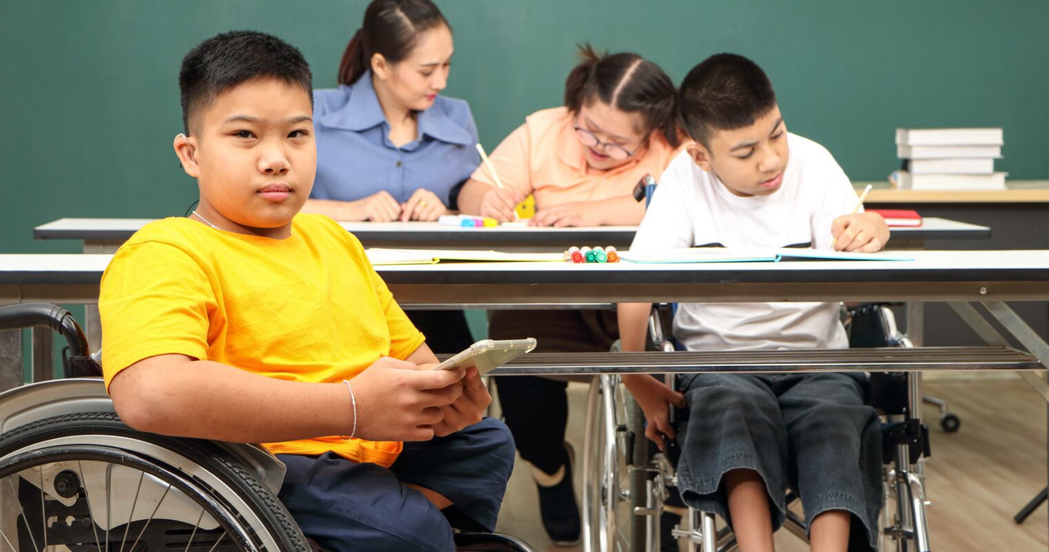 A student wearing an orange shirt, sitting in a wheelchair, looking at the camera, while other students work at desks in the background.