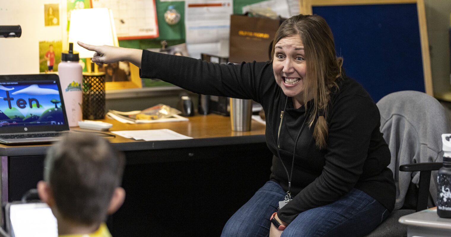 A teacher wearing a black shirt pointing to something, while happily working with a student in a classroom.