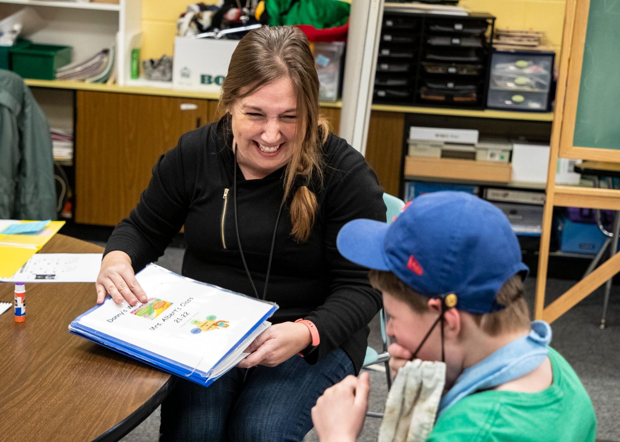 In a classroom, a teacher in a black shirt, laugh with a student while showing them educational materials.