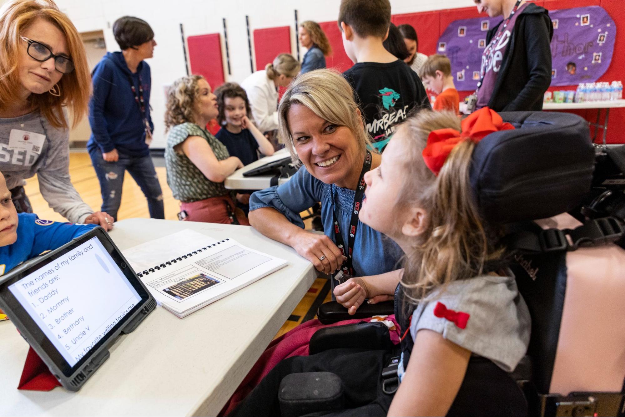 A special education teacher smiles with a student working on an AAC device.
