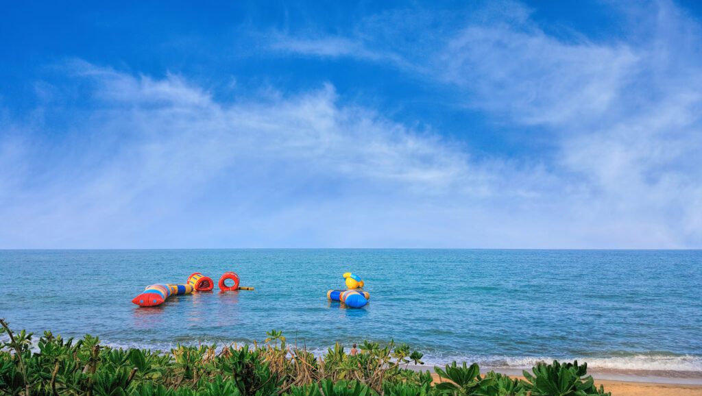 A beach scene with open water and a floating trampoline anchored off the shore in an image with blue skies and wispy clouds