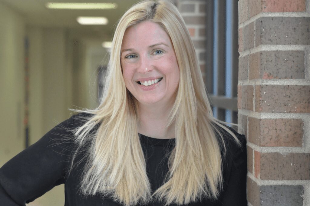Woman with long blonde hair smiling toward the camera in a school hallway.