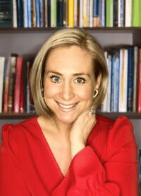 Headshot of a woman (Lauren Sheehan) with shoulder-length blonde hair wearing a red top with a bookcase in the background.