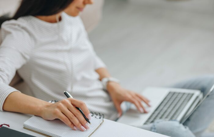 A woman learning remotely with one hand on the keyboard of a laptop computer and another hand holding a pen near a pad of paper.