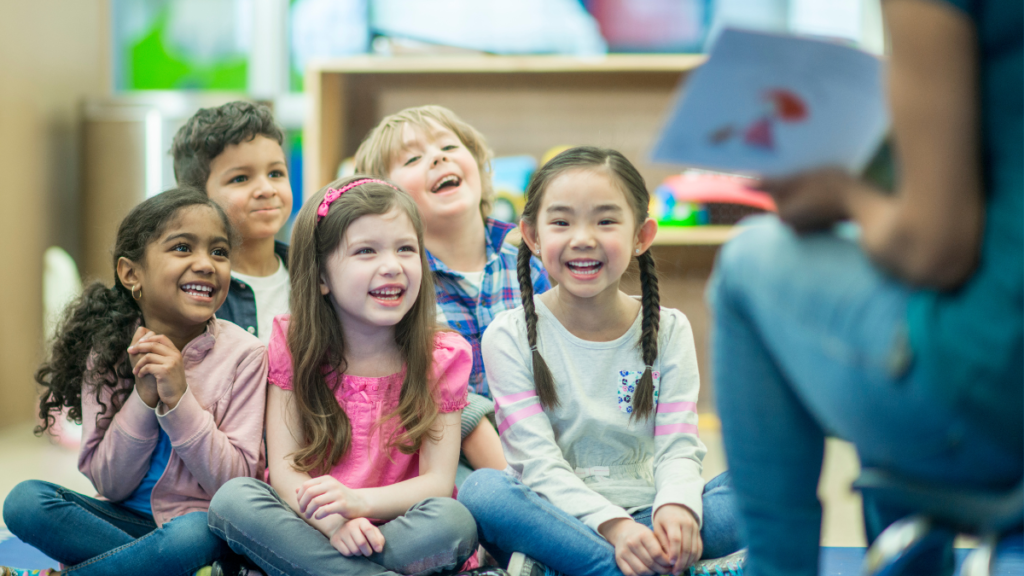 A group of young students sitting on a classroom floor facing a teacher who is reading out loud from a book.