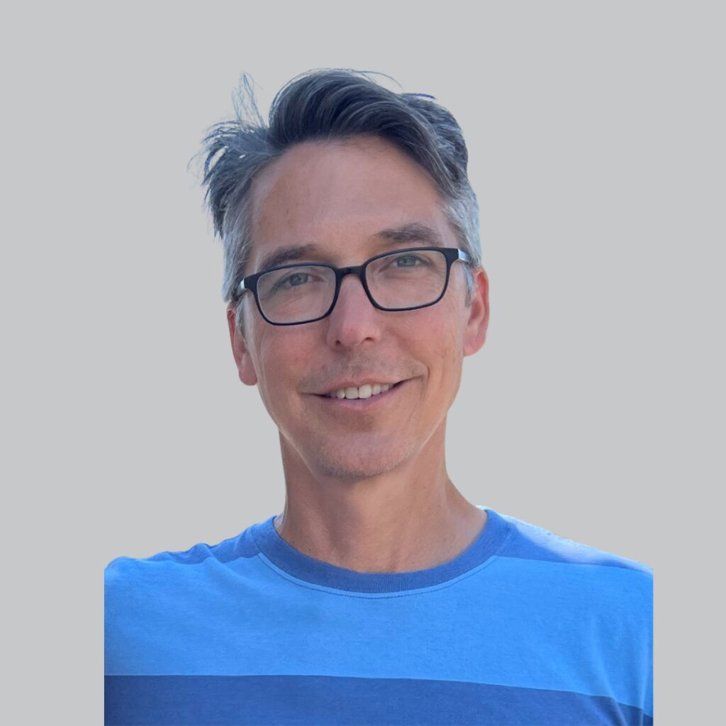 man with grey hair and blue shirt smiles in front of a grey background