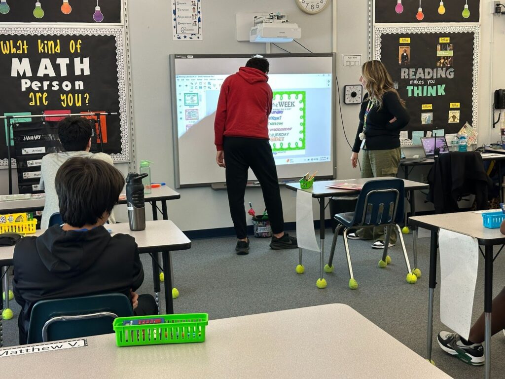 Learner provides specially designed instruction in a red hoodie at the whiteboard in front of class.