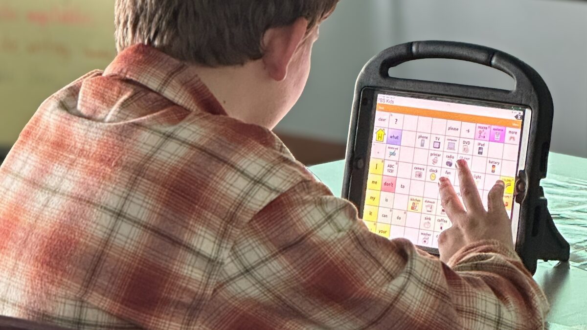 Child wearing plaid looks at their AAC device to learn and communicate