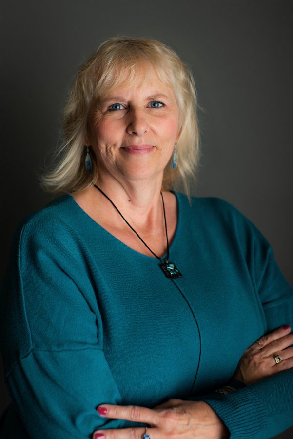 Woman with blonde hair and teal shirt smiles in front of a dark grey background.