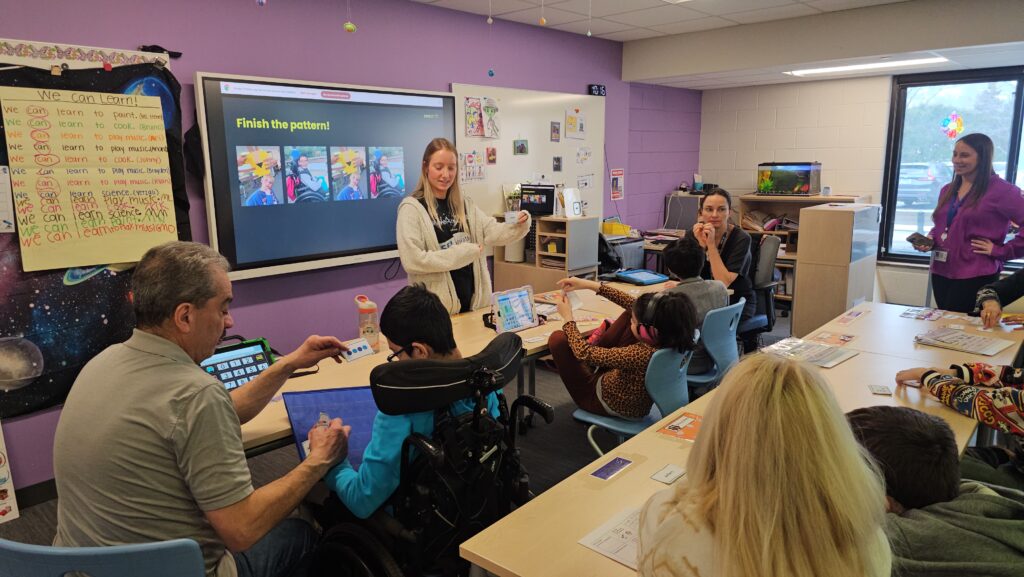 Teacher leads a hands-on activity at a table while students use tablets and sorting cards in a purple-walled classroom.