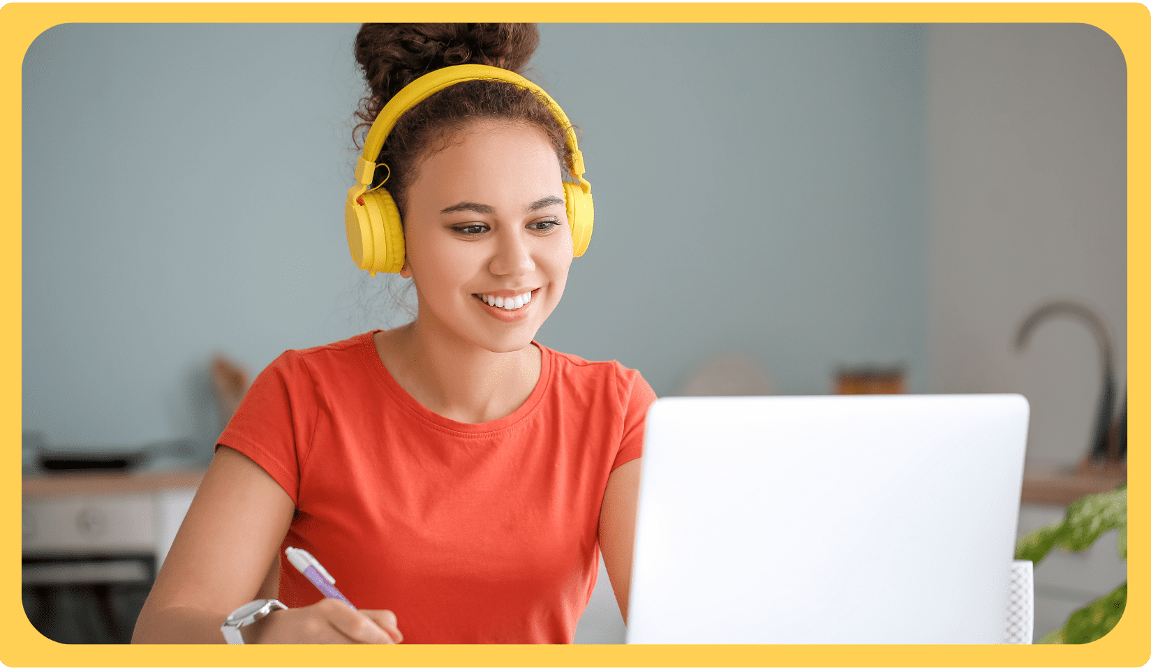 Girl wearing bright yellow headphones smiles at a laptop while writing in a notebook in a cozy room.