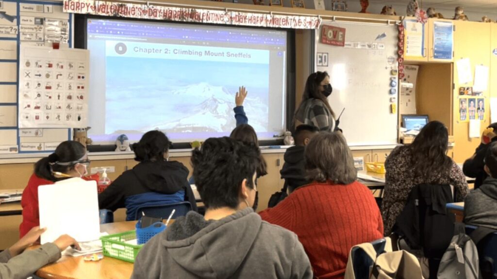 Classroom scene with a masked teacher at the whiteboard and students seated, watching a projection that reads 'Chapter 2: Climbing Mount Sneffels'. Valentine banner overhead.