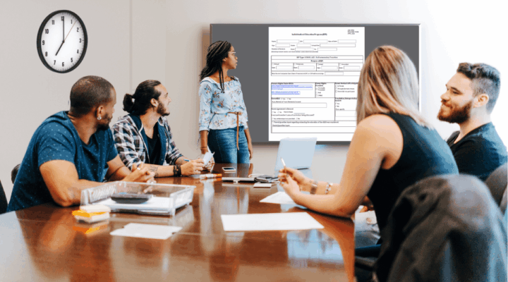 Diverse team around a conference table watching a presenter with a form displayed on a wall screen behind her.