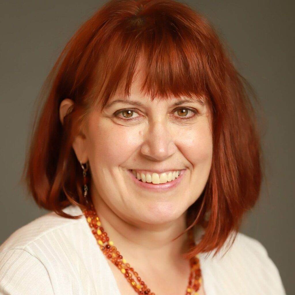 Close-up portrait of a smiling woman with shoulder-length red hair and brown eyes, wearing a white top and orange bead necklace.