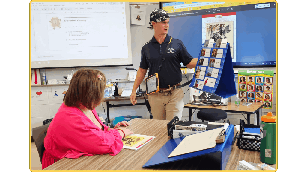 Male teacher in a navy polo and bandana stands beside a blue card display, presenting to a seated student in a pink top in a bright classroom.