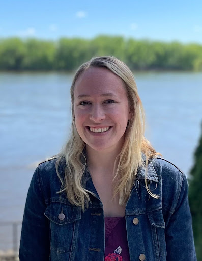 Woman wearing a denim jacket over a red shirt with blonde hair smiles in front of a background of water and trees.